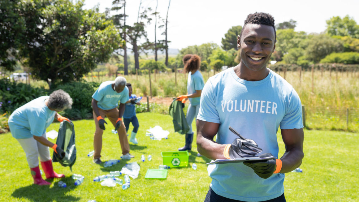 Portrait of a smiling young African American man standing in a field wearing wearing a t shirt with volunteer written on it and gloves, writing on a clipboard and smiling to camera, while a diverse group of volunteers collect rubbish and recycling in the b Helping People Who Need It